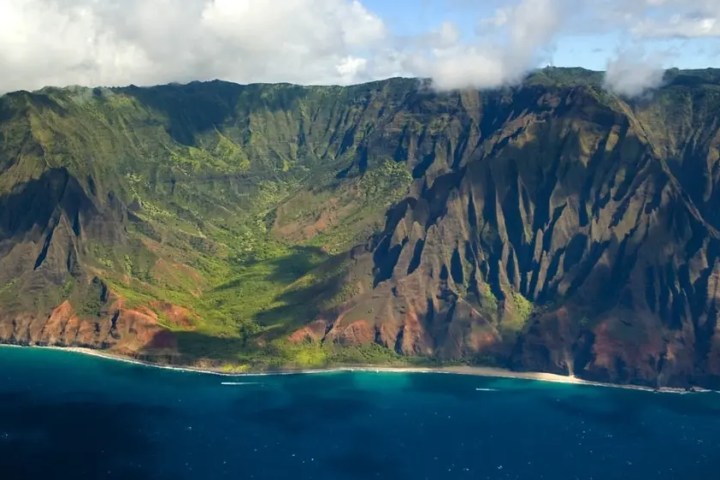 Aerial view of a lush green coastline with steep cliffs and blue ocean under cloudy skies.