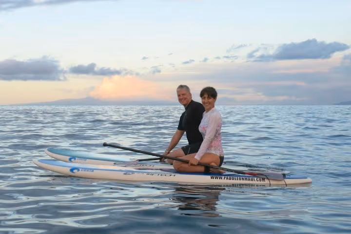 Two people sitting on paddleboards on a calm ocean at sunset.