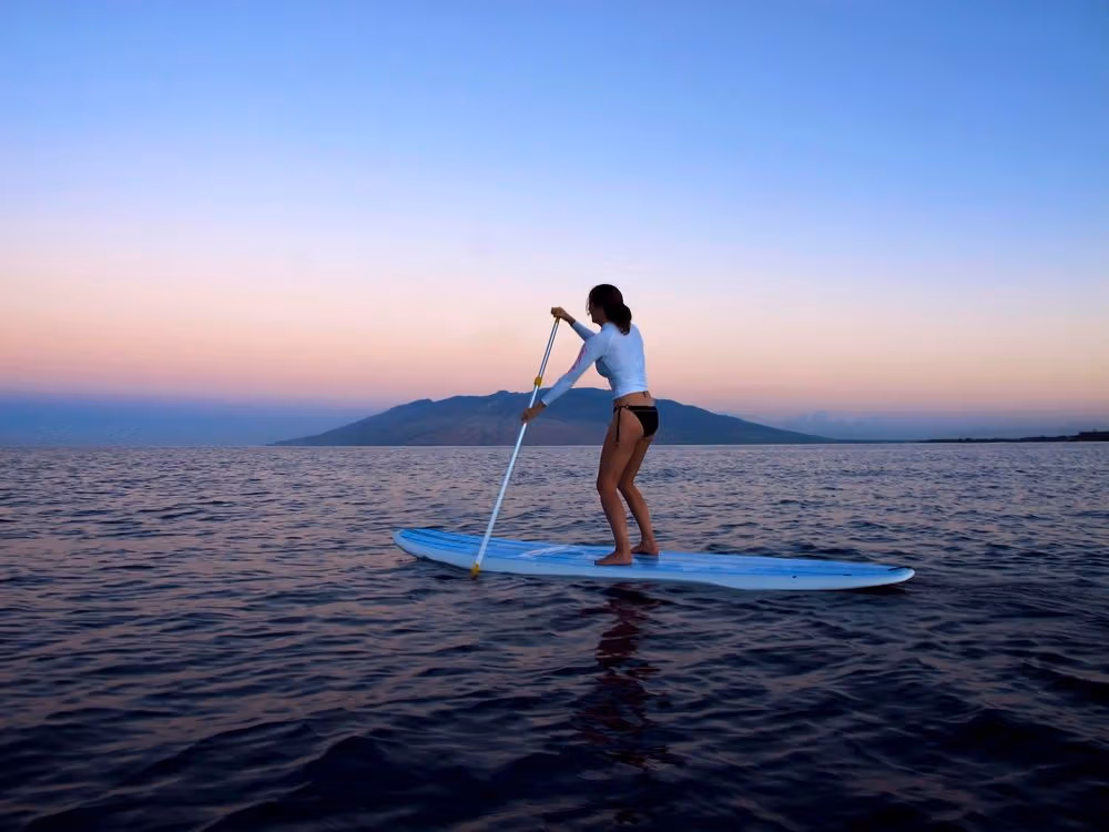 Person paddleboarding on calm sea at sunset with mountain in background.