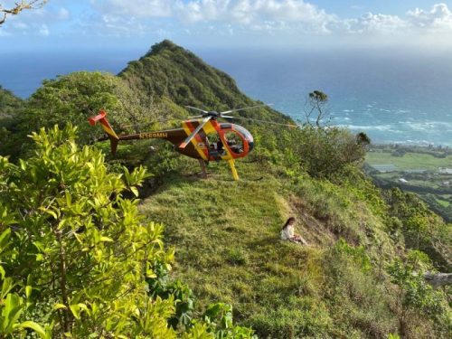 Kualoa-Ranch-Ridgeline-LZ Red and yellow helicopter lands on a grassy hilltop near the ocean with a distant mountain view.