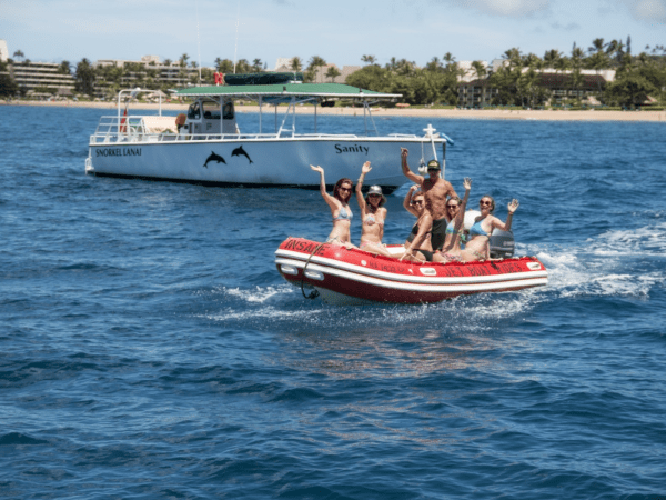 People waving on a red inflatable boat with a tour boat named Sanity in the background on the ocean.