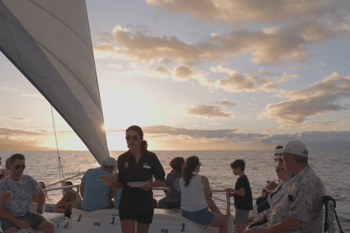 Group of people on a sailboat at sunset with a woman serving drinks.