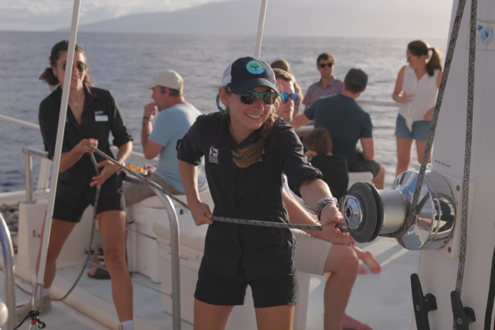 Woman operating a winch on a boat with others gathered around on deck.