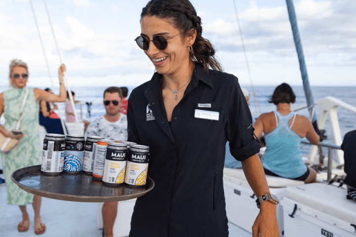 Smiling woman on a boat carrying a tray of assorted canned drinks with people in the background.