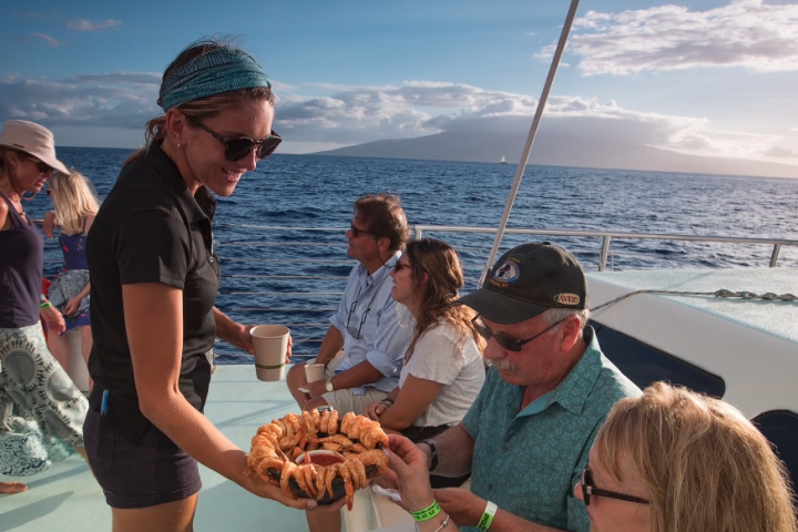 People on a boat enjoying snacks and drinks with a scenic ocean view at sunset.