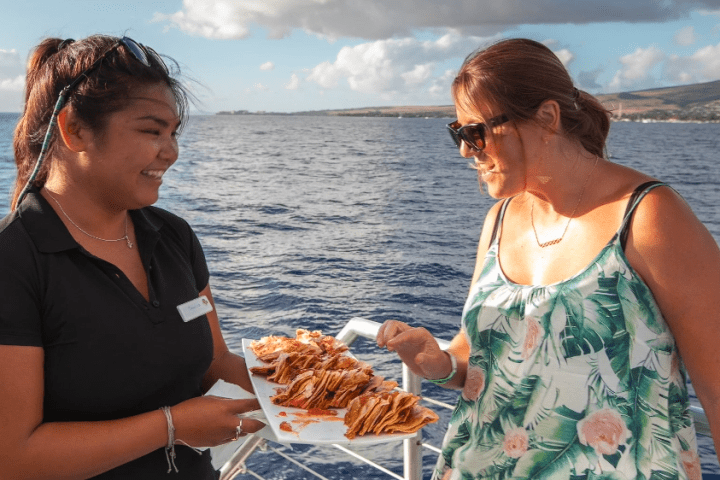 Two women on a boat with a plate of food, one serving the other under a sunny sky.