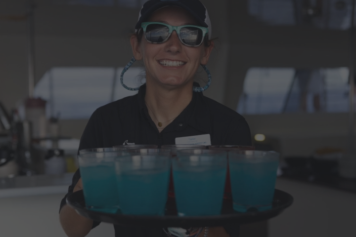 Smiling person wearing sunglasses holding a tray of blue drinks indoors.