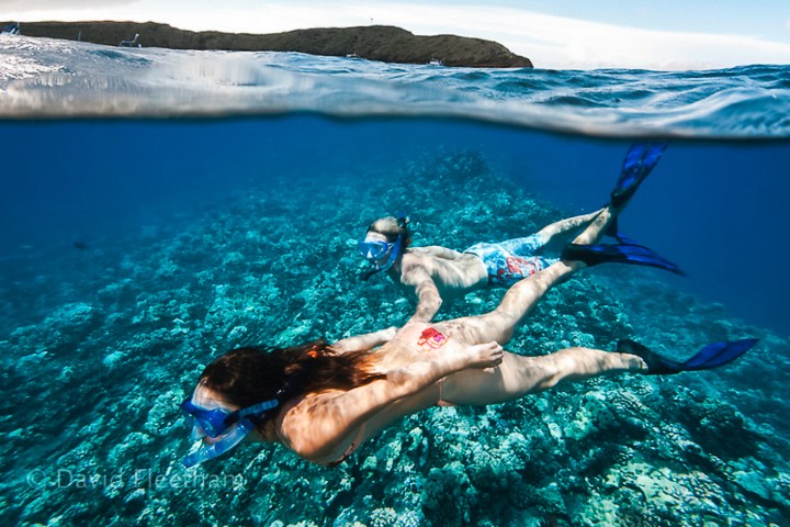 Two snorkelers swimming over coral reef with clear water and hills in the background.