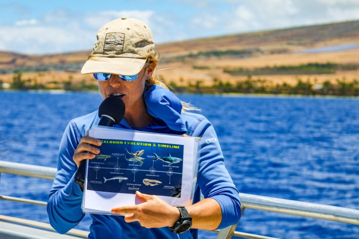 Person in blue shirt holds a microphone and timeline chart near a body of water.