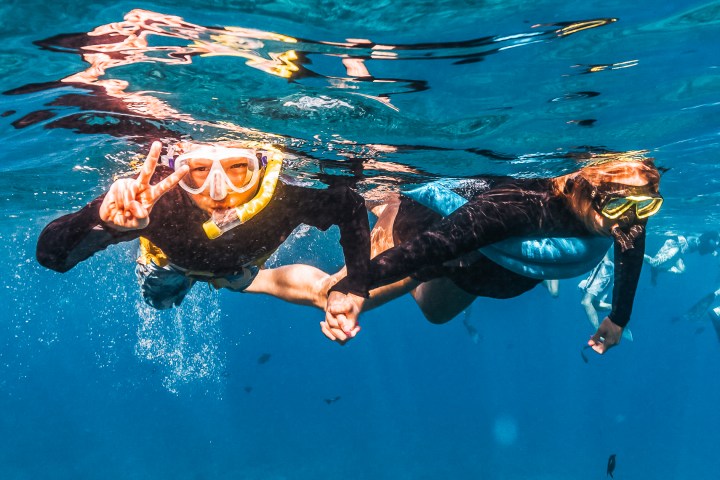 Two people snorkeling underwater, holding hands and wearing masks in clear blue water.