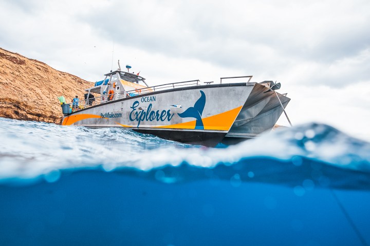 Boat named Ocean Explorer on water near rocky shore with cloudy sky.