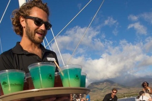 Man with sunglasses holds a tray of blue-green drinks outdoors with people and mountains in the background.