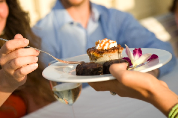 Hands offering dessert plate with smiling people in background.