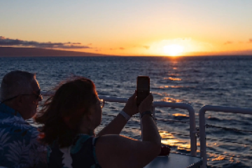 Two people on a boat capturing a sunset over the ocean with a smartphone.