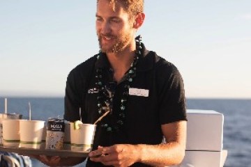 Man in black shirt serving drinks on a tray by the ocean at sunset.