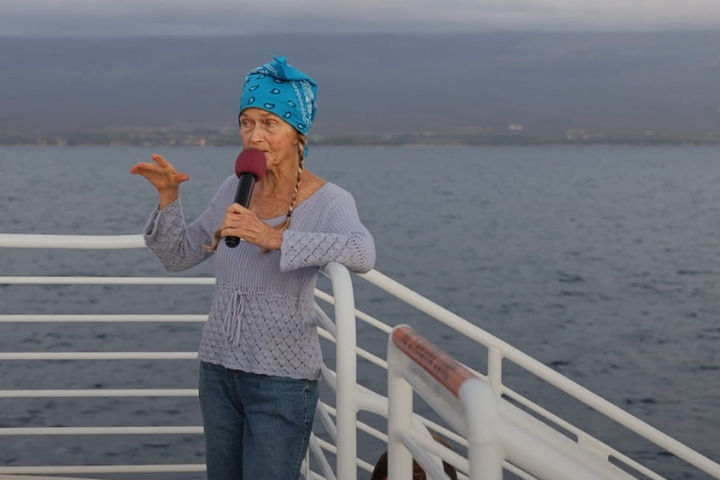 Person with blue bandana speaking into microphone on a boat deck, ocean in the background.