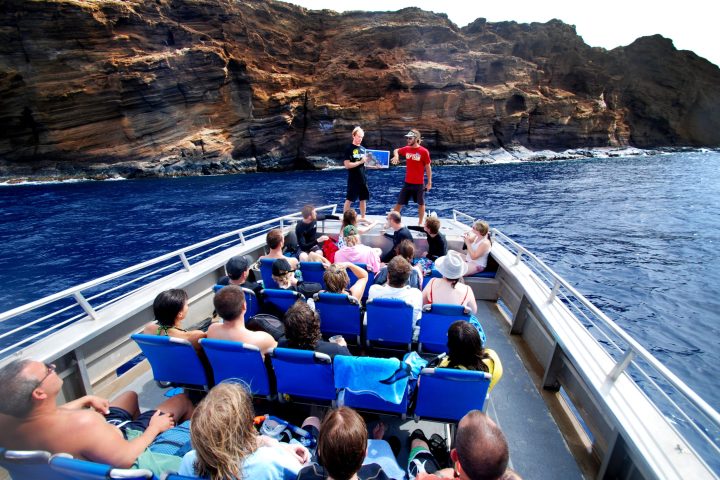 Tourists on a boat with blue seats near a rocky cliff by the ocean.