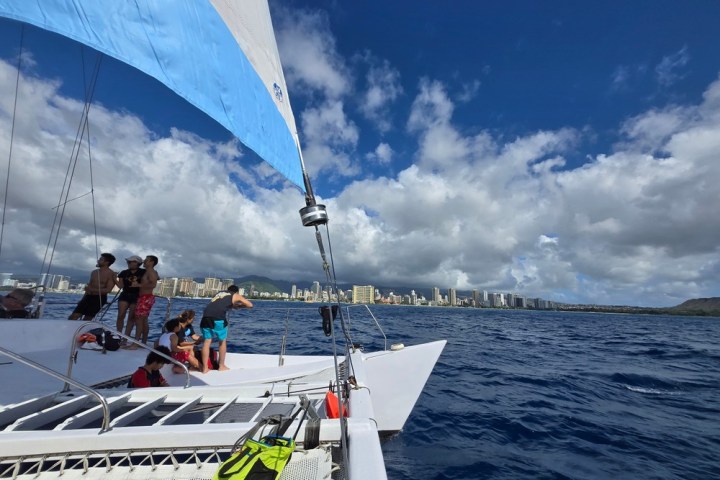 People on a sailboat with city skyline and cloudy sky in the background.