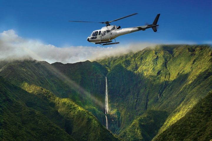 Helicopter flying over lush green mountains with a waterfall in the background.