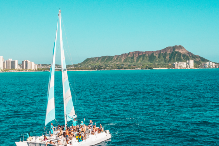 Catamaran with people on blue ocean, city skyscrapers, and mountain in background under clear sky.