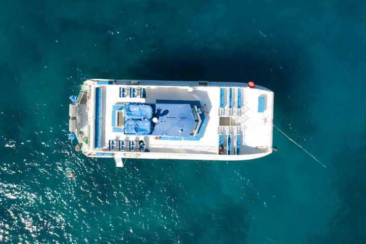 Aerial view of a white catamaran on blue ocean, with empty deck chairs and tarps.