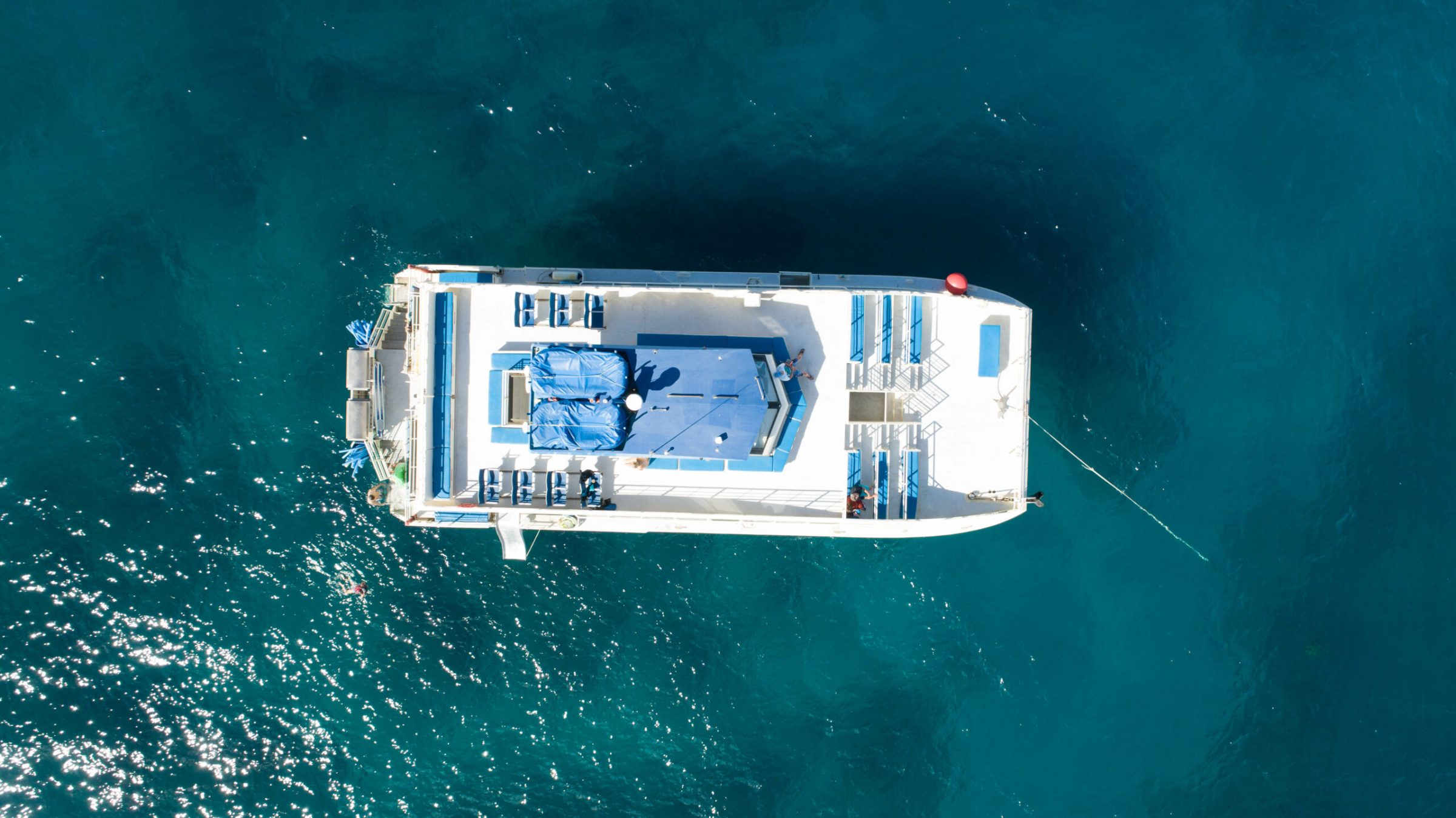 OV_Top-view-scaled Aerial view of a white catamaran on blue ocean, with empty deck chairs and tarps.