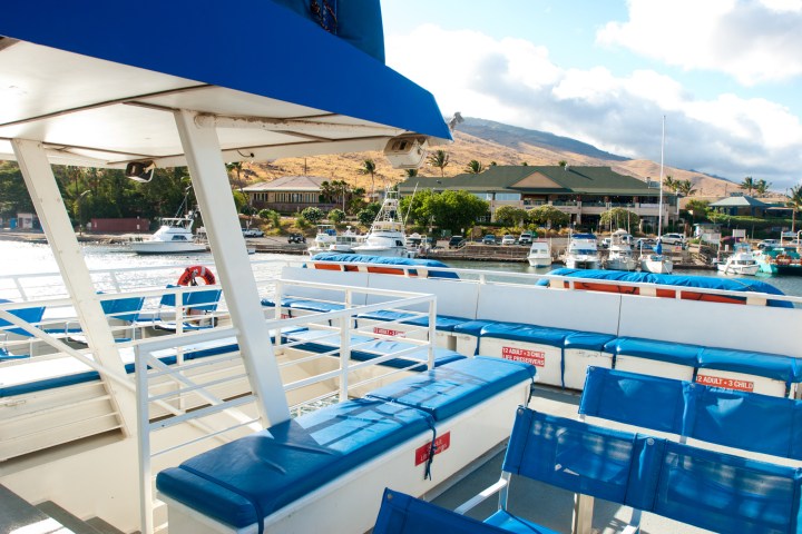 View from a boat with blue seats, facing a marina with boats and hills under a cloudy sky.
