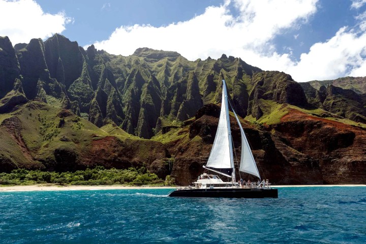 Sailboat on blue water with dramatic green cliffs and a partly cloudy sky in the background.
