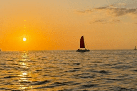 Sailboat with red sail on ocean at sunset, orange sky and clouds.