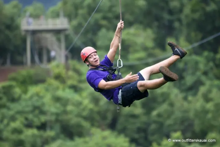 Person ziplining through forest wearing helmet, purple shirt, and shorts.