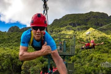 Two people ziplining in helmets over lush green landscape and mountains.