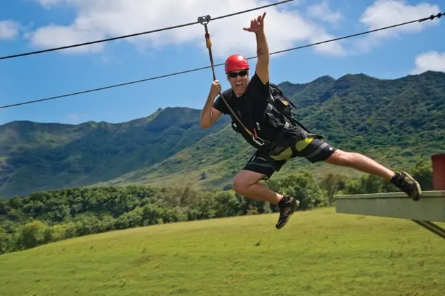 Person zip-lining with a red helmet and sunglasses, against a background of mountains and clear sky.