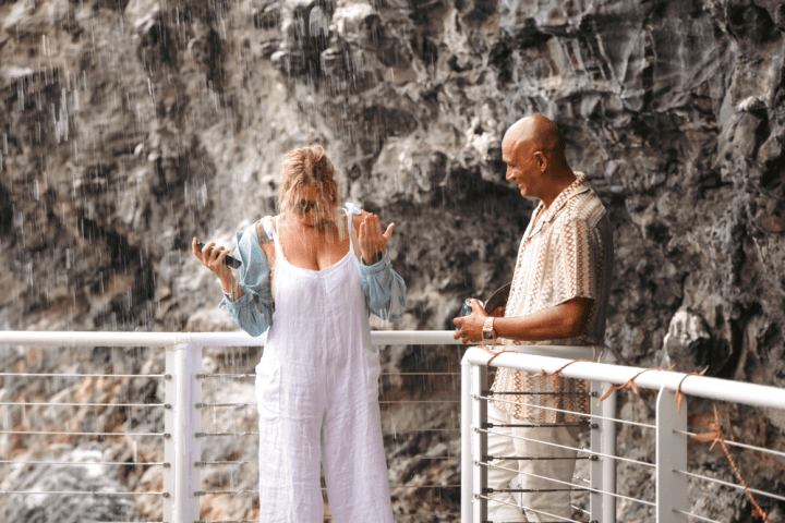 Woman standing in rain on deck, smiling man beside her against rocky background.
