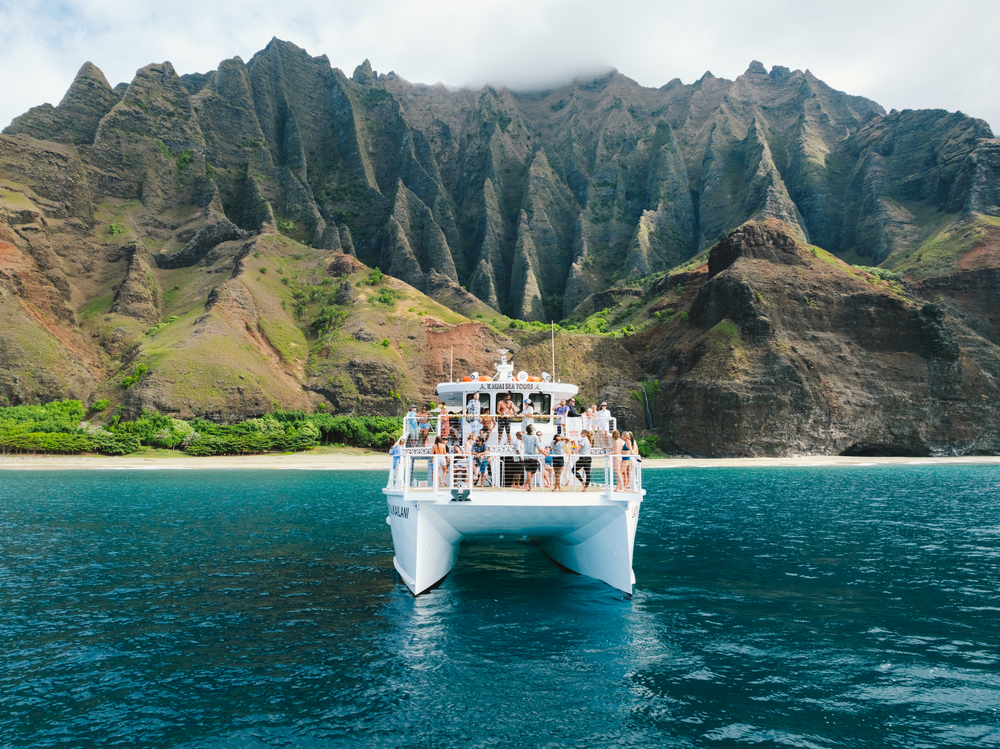 Boat with tourists in front of rugged, green cliffs and blue water.
