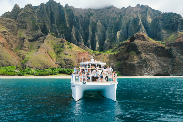 Boat with tourists in front of rugged, green cliffs and blue water.