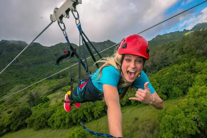 Person zip-lining over forested area with mountains, wearing a red helmet and blue shirt, smiling and gesturing.