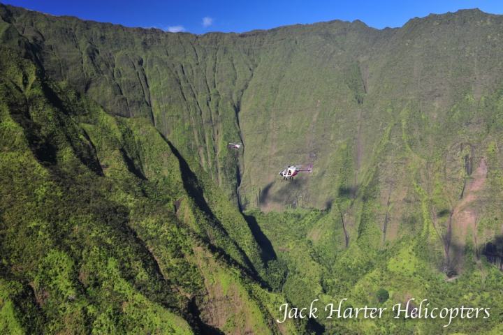 Helicopter flying over lush green cliffs under a clear blue sky.