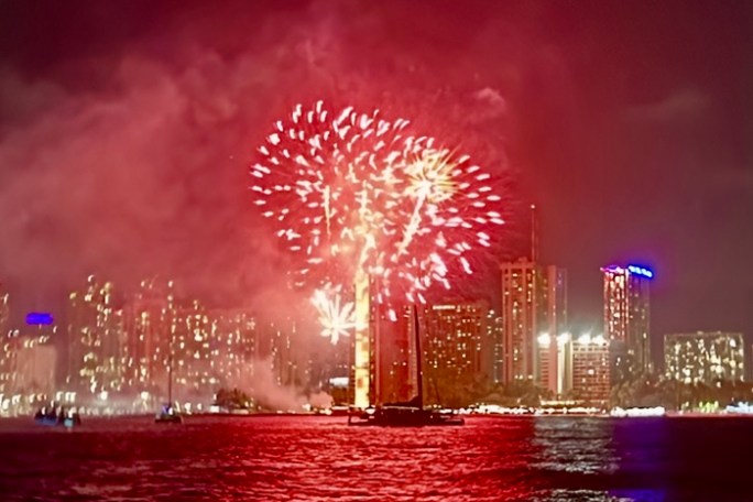 Fireworks over a cityscape with red reflections on water at night.