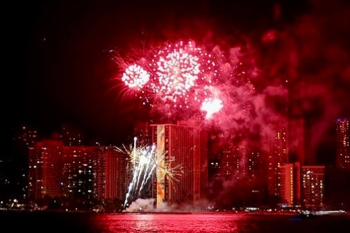 Red fireworks over a city skyline reflecting on water at night.