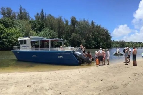 People gather near a blue boat on a sandy riverbank under a clear sky.