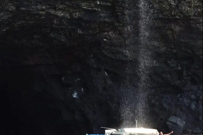 A boat near a rock wall with a waterfall, and people on the deck enjoying the view.