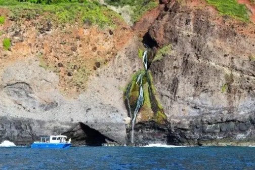 Blue boat near rocky cliff with waterfall and green vegetation above.