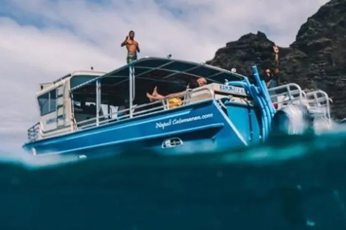 Blue tour boat with people on deck, ocean water in foreground, rocky cliffs in background.