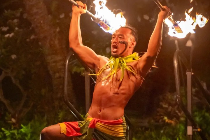 Performer kneeling on stage at night, holding flaming torches, wearing traditional attire, facial expression intense.