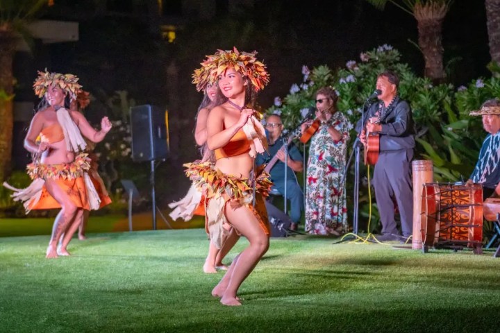 Hula dancers in traditional attire perform on grass with musicians playing in the background.