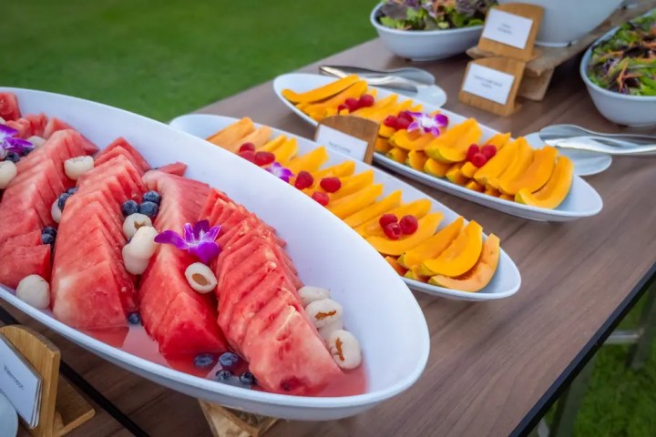 Plates of sliced watermelon, papaya, and berries on a wooden table outdoors.