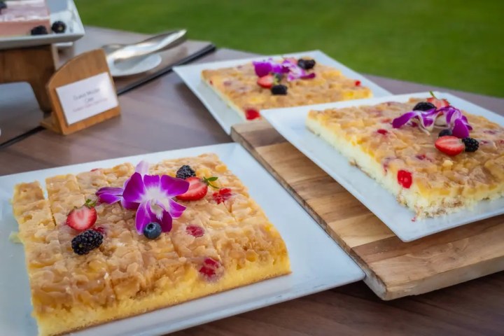 Fruit-topped sheet cakes with flowers on white plates, displayed outdoors on a wooden table.