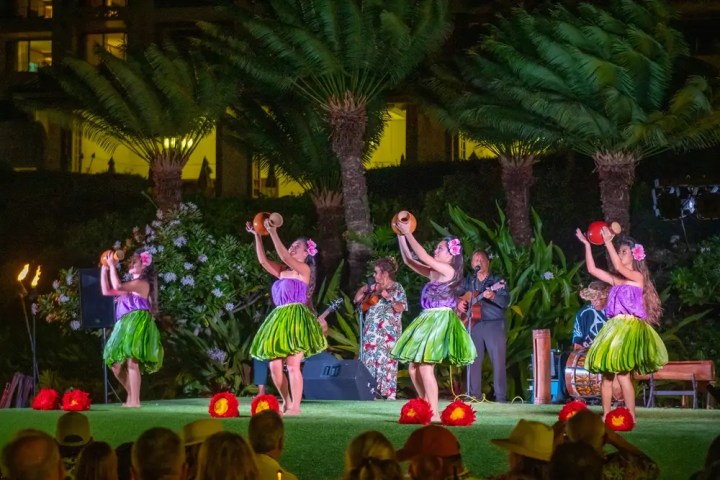 Four women in colorful Hawaiian attire dance on stage, with musicians in the background under palm trees.