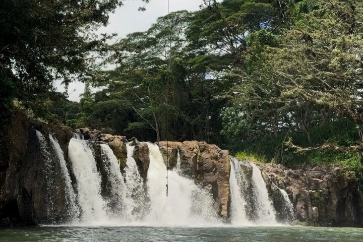 Waterfall cascading into a river, surrounded by lush greenery under a partly cloudy sky.