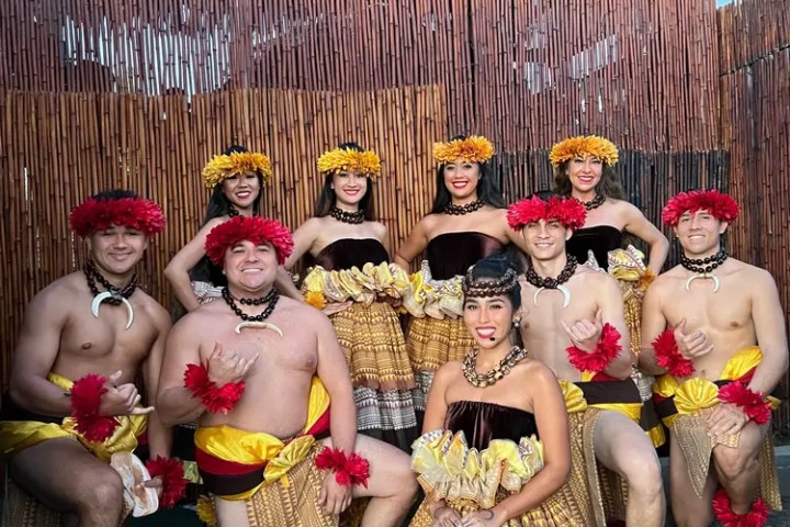 Group in traditional Hawaiian attire posing against a bamboo wall.